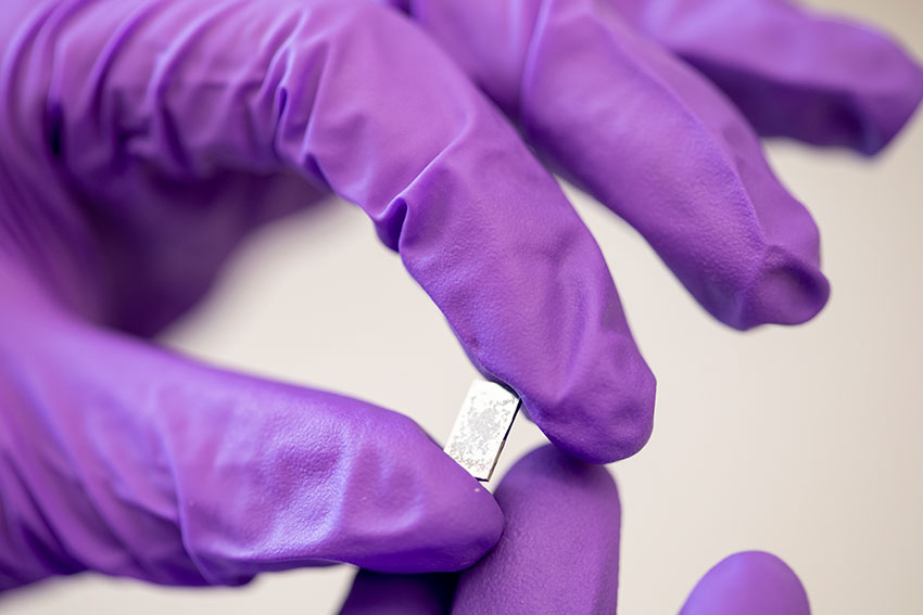 Hands in purple lab gloves hold a small silver wafer.