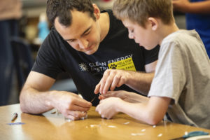 A young man with a black t-shirt and a yellow nametag sits at a table with a young student. The two are working together to assemble a small solar spinner from a petri dish.