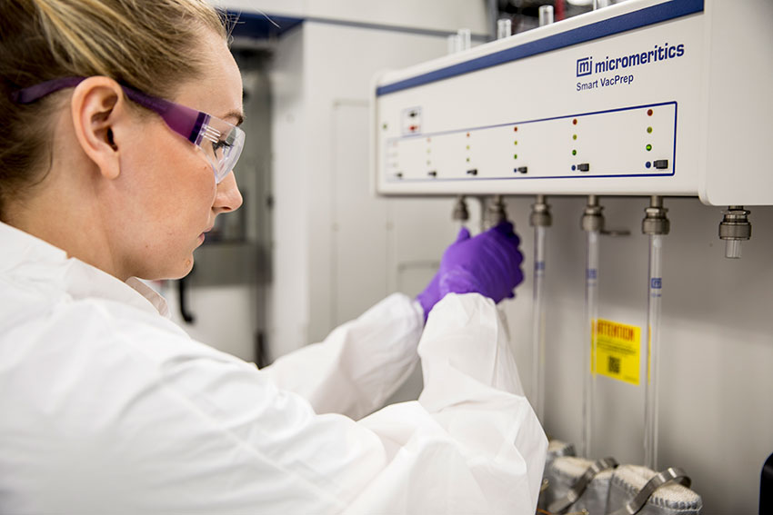A young woman in a lab coat, protective glasses, and lab gloves adjusts the settings on a piece of scientific equipment.
