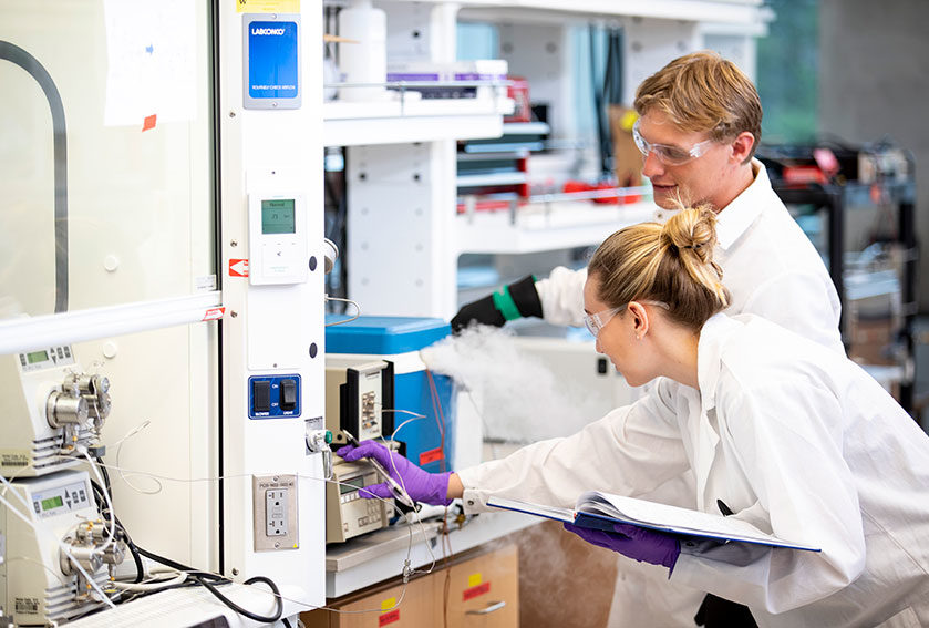 A young woman and man in lab coats and protective glasses operating lab machinery.