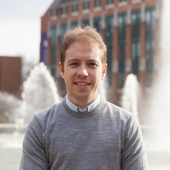 A man in a collared shirt and sweater poses in front of a fountain.