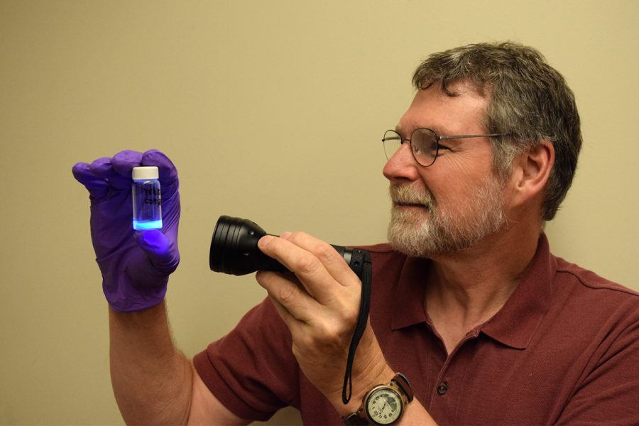 A man wearing glasses shines an ultraviolet flashlight onto a small container; the contents glow blue.