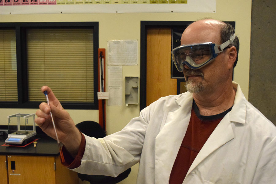 A man in a lab coat and goggles holds a test tube containing a red liquid.