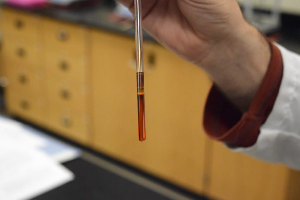 A hand holds a small test tube containing a clear red liquid.