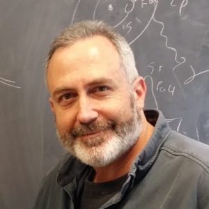 A headshot of a man standing in front of a chalkboard.