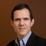 A middle-aged man in a jacket and collared shirt looks confidently into the camera.