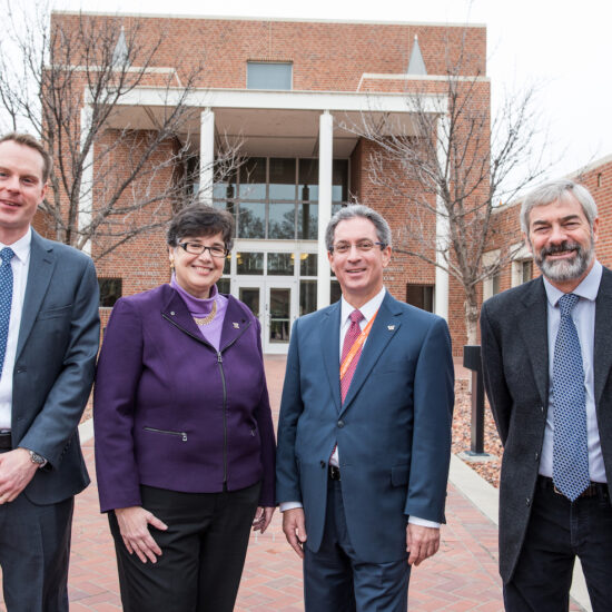 A woman in a purple jacket and sweater and three men in suits pose in front of a brick building.