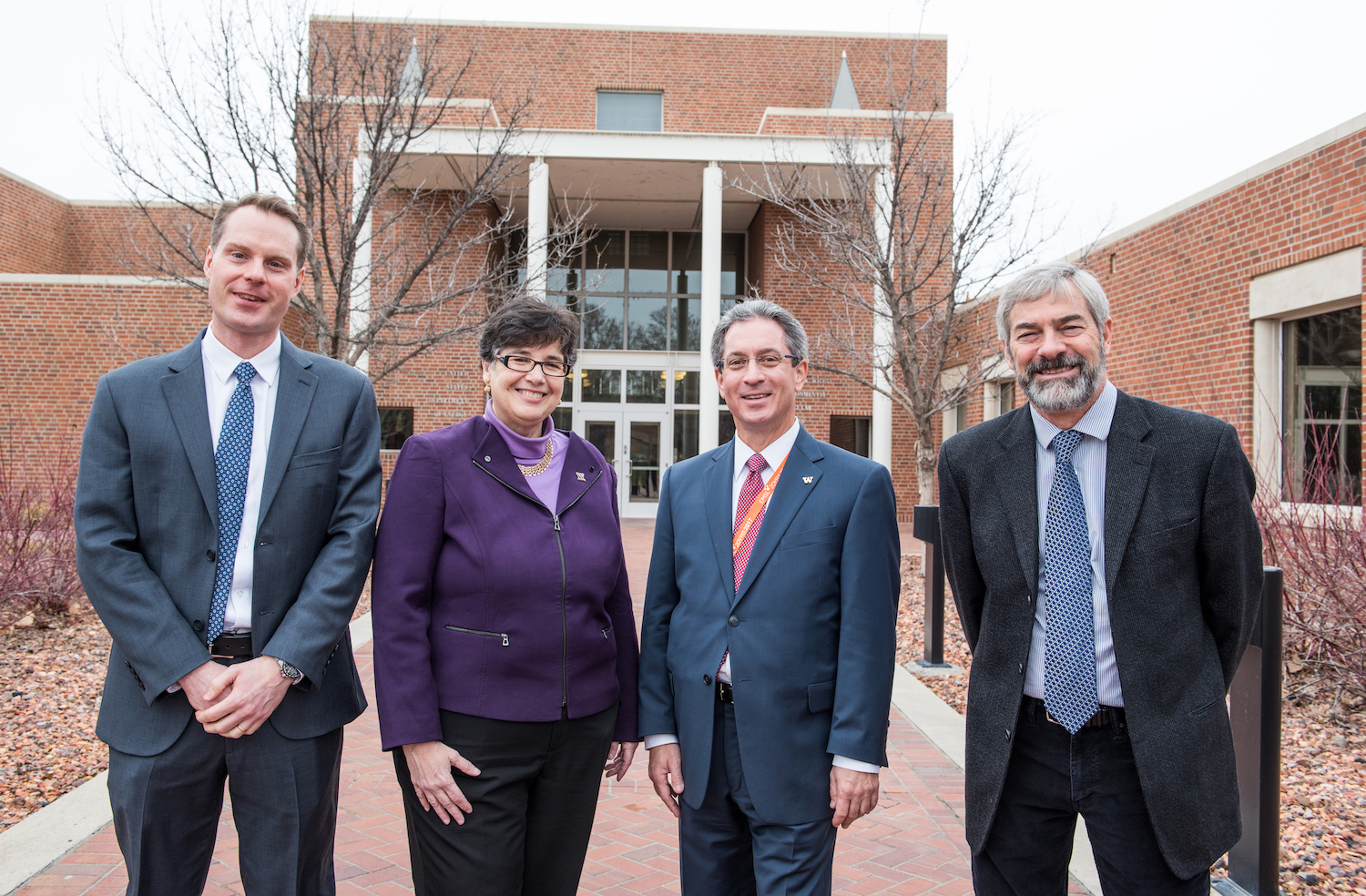 A woman in a purple jacket and sweater and three men in suits pose in front of a brick building.