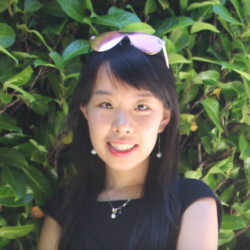 A young woman with sunglasses on top of her head smiles for the camera in front of a leafy background.