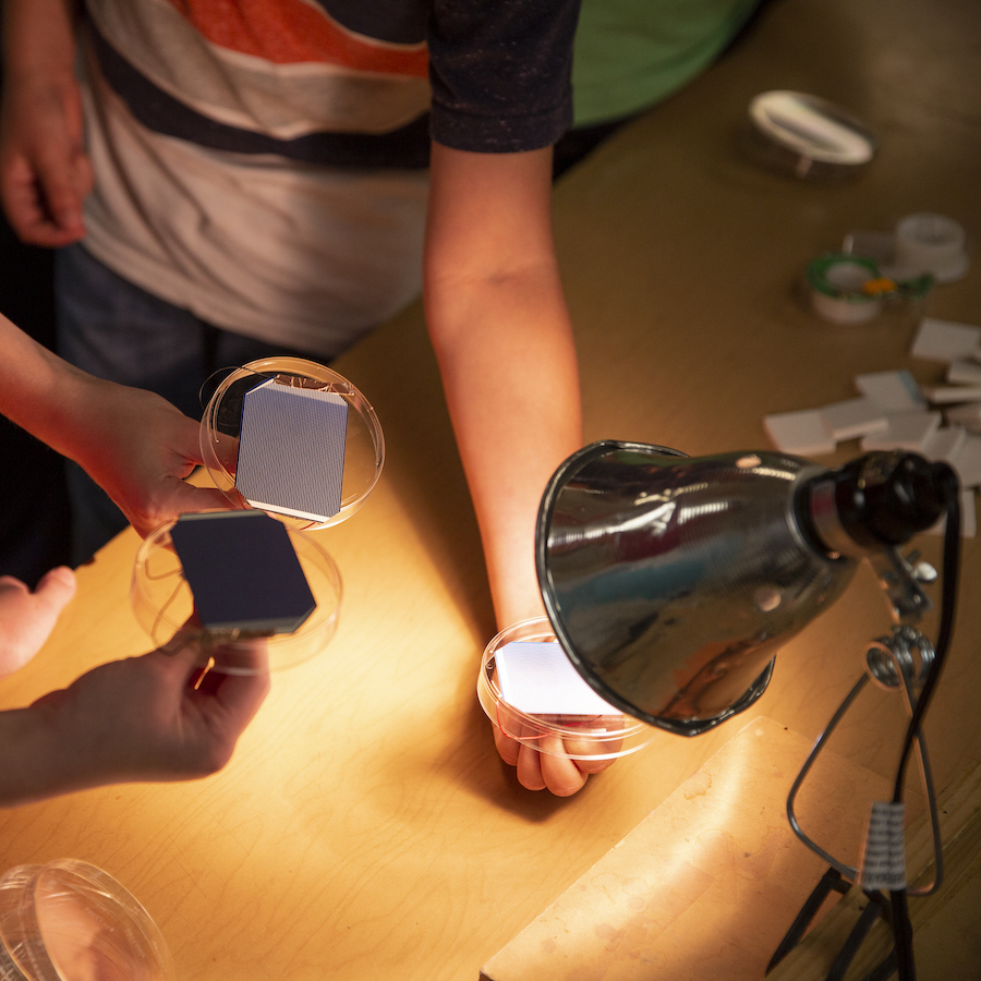 Two students hold their solar spinners - solar panels glued to petri dish lids - underneath a high-intensity lamp.