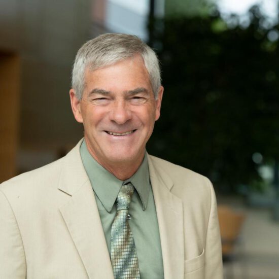 Jim De Yoreo smiles for the camera wearing a tan suit, sage green shirt, and patterned green tie.